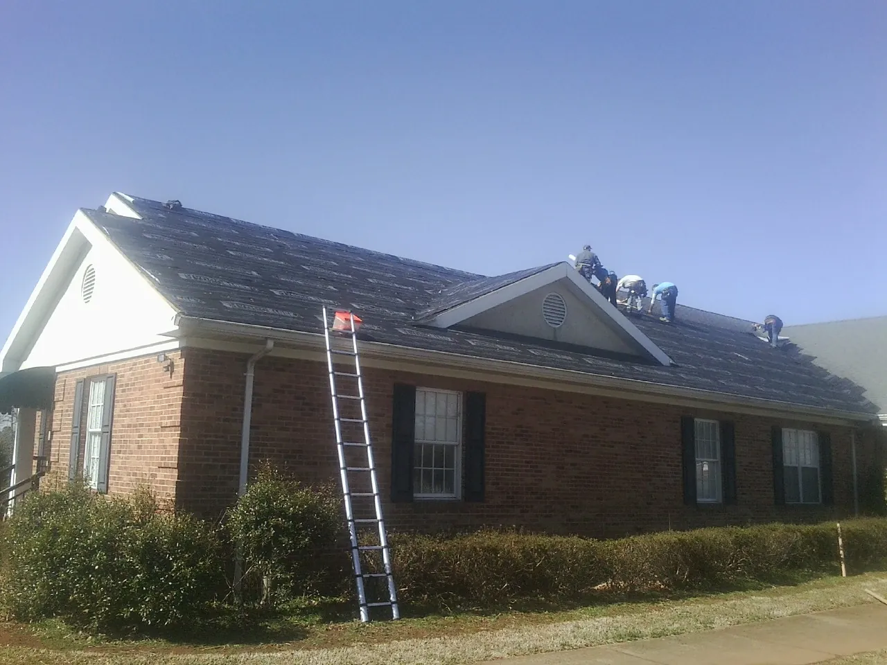 Crew tearing shingles off a brick ranch roof with a ladder set — Emergency Roof Repair work by our crew in Newfane